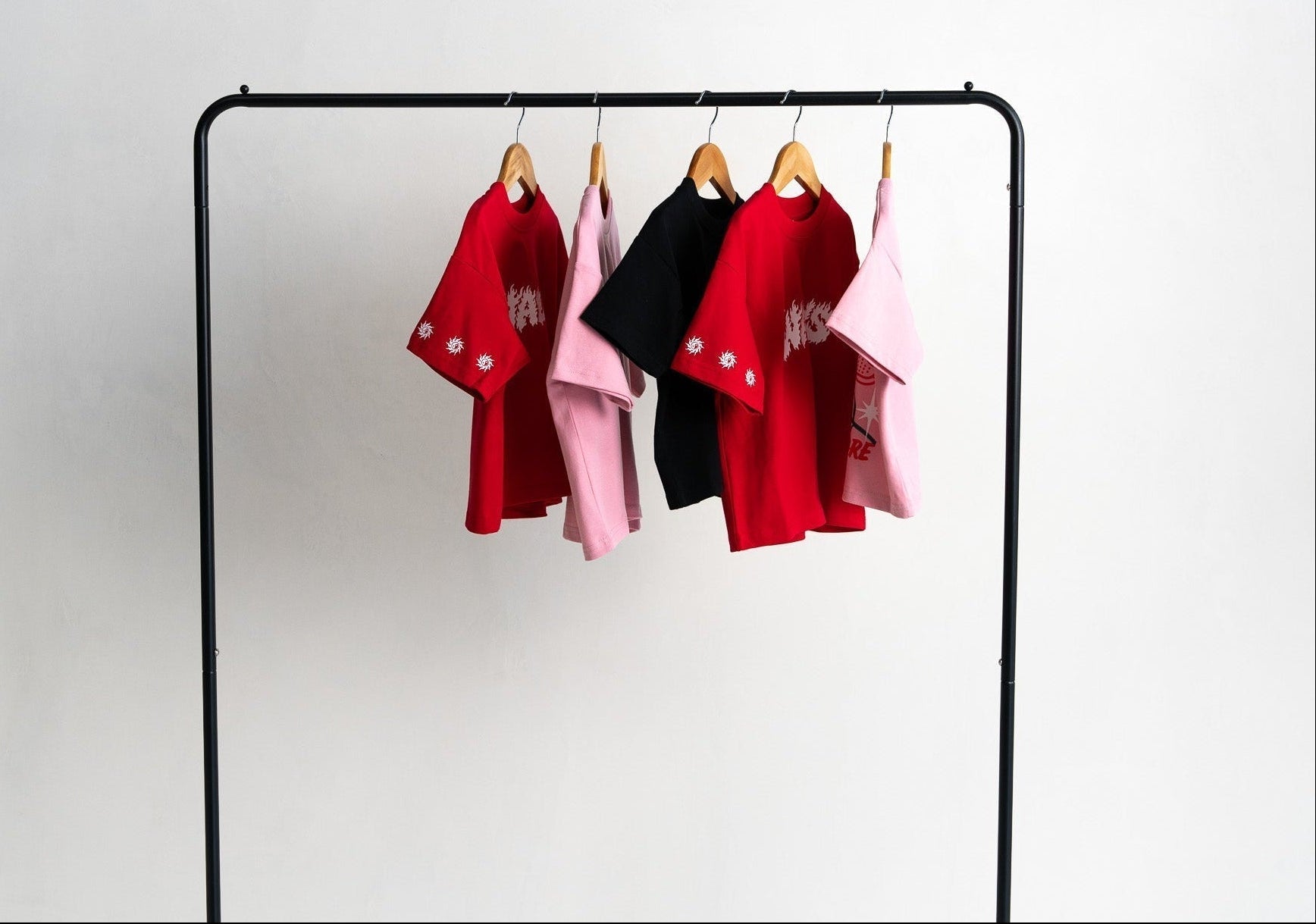 Four short-sleeve t-shirts in red, black, and pink hanging on wooden hangers from a black metal clothing rack against a plain white wall.
