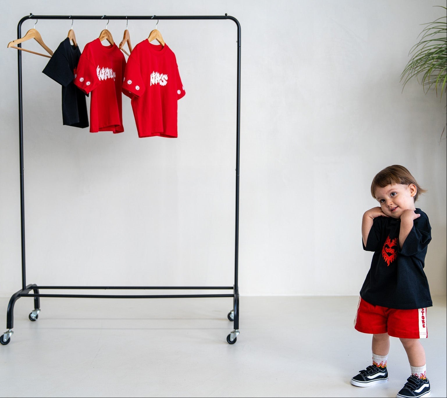A toddler stands on the right wearing a black graphic T-shirt and red shorts, next to a black rolling clothing rack with two red T-shirts and one black T-shirt hanging on wooden hangers against a white wall.