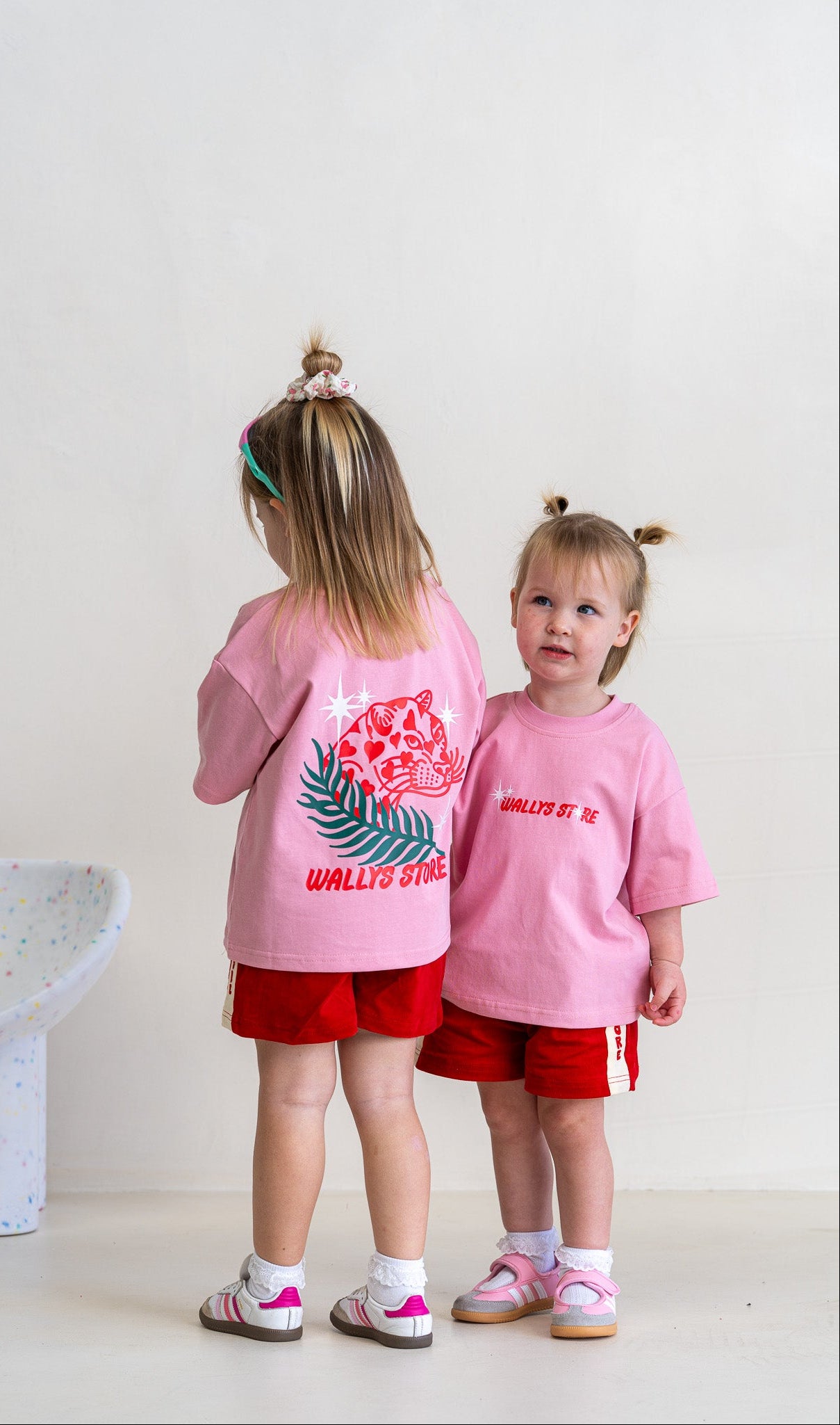 Two young girls modeling pink t-shirts with red shorts in a bright, minimalist room. The girl on the left is shown from the back, wearing a pink shirt with a large red and green tiger graphic and the text 'WALLIS ST.