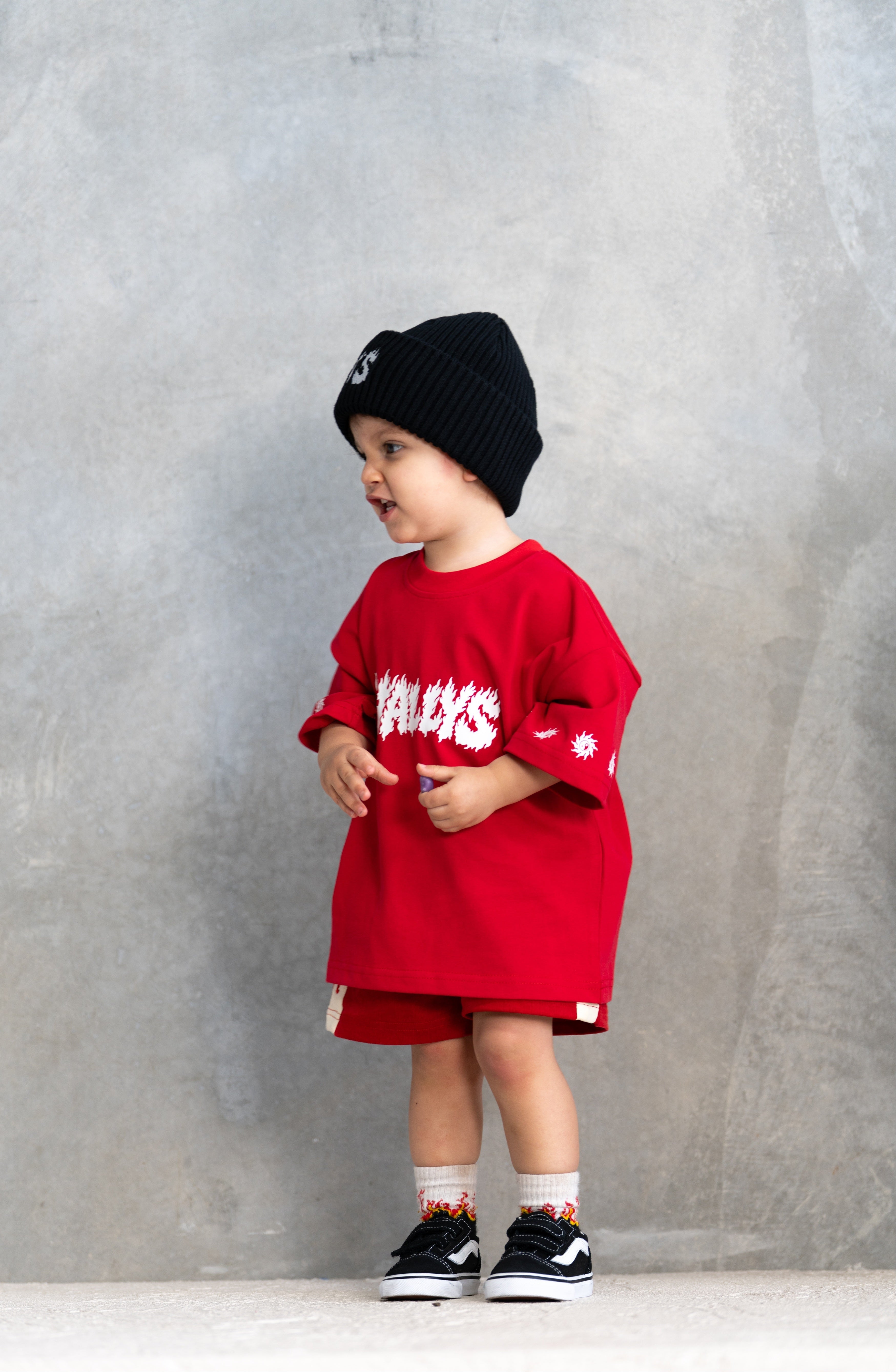 A toddler stands against a textured gray wall wearing an oversized bright red graphic T-shirt dress with white lettering, matching red shorts, a black knit beanie, white socks, and black sneakers.