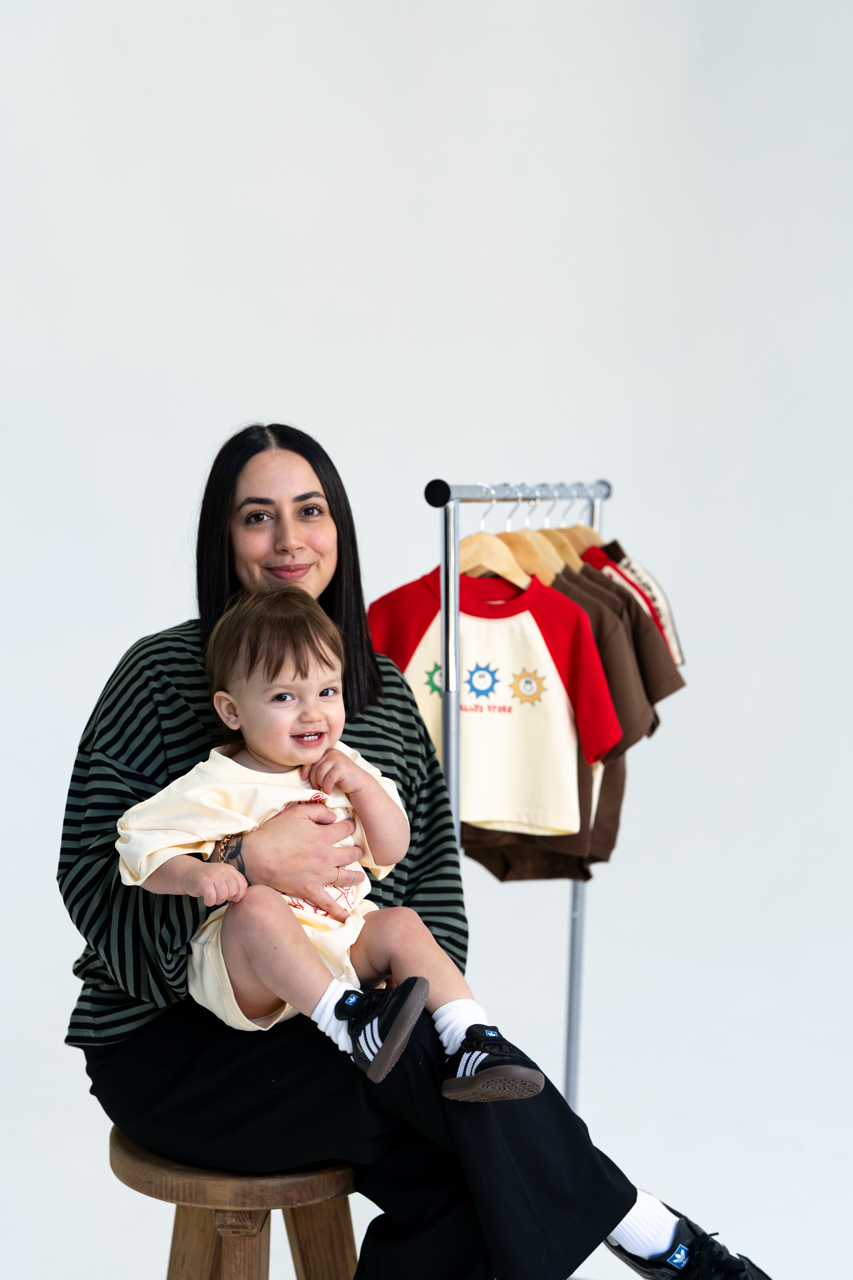 Woman holding a child sitting on a stool with a rack of clothes in the background Wallys Store Kids Clothing